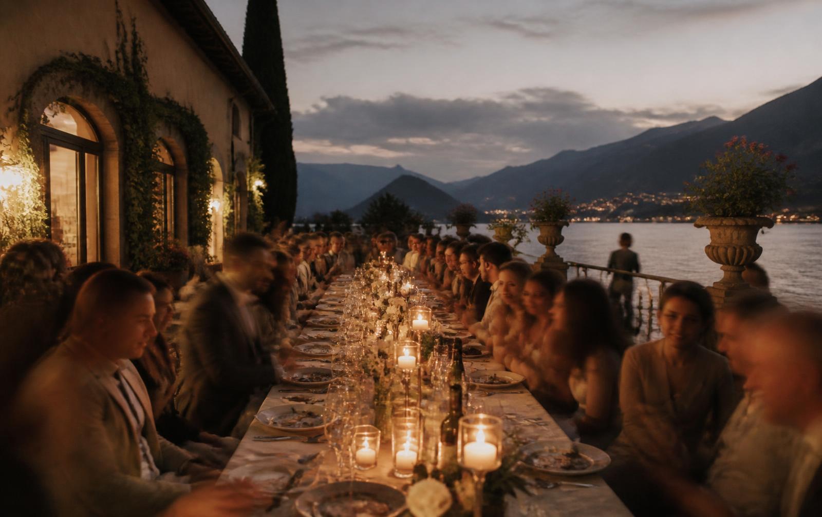 Long candlelit banquet table on a terrace above the lake at dusk